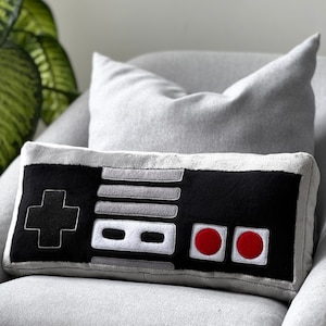 May include: A black and grey throw pillow shaped like a classic video game controller. The pillow has a white plus sign, two white buttons, and two red buttons.