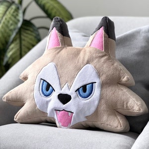 Könnte beinhalten: Ein Plüschkissen in Form eines Wolfskopfes mit weißem Gesicht, blauen Augen und einer herausgestreckten rosa Zunge. Das Kissen ist beige mit schwarzen Ohren und rosa Akzenten.