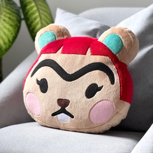 Op de afbeelding: Een pluche kussen in de vorm van een cartoonfiguur met rood haar, een bruine neus en een witte tand. Het personage heeft een zwarte wenkbrauw en grote roze wangen.