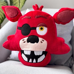 May include: Red plush pillow shaped like the head of a cartoon fox with a black eye patch and one yellow eye. The fox has a wide smile with white teeth.