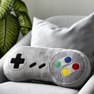 May include: A gray plush pillow shaped like a classic video game controller. The pillow has a black plus sign, four black dots, and four colored buttons: green, blue, red, and yellow.