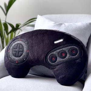 May include: A plush pillow shaped like a black video game controller. The controller features a circular directional pad with grey accents, three circular buttons labeled A, B, and C in red, and a white rectangular button on the upper right side.