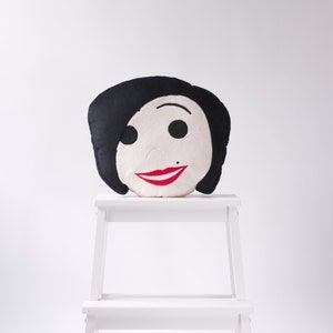 May include: A decorative pillow shaped like a face with black hair, black eyes, a small beauty mark, and a red smile. The pillow is placed on a small white step stool against a white background. The pillow's design is simple and cartoon-like.