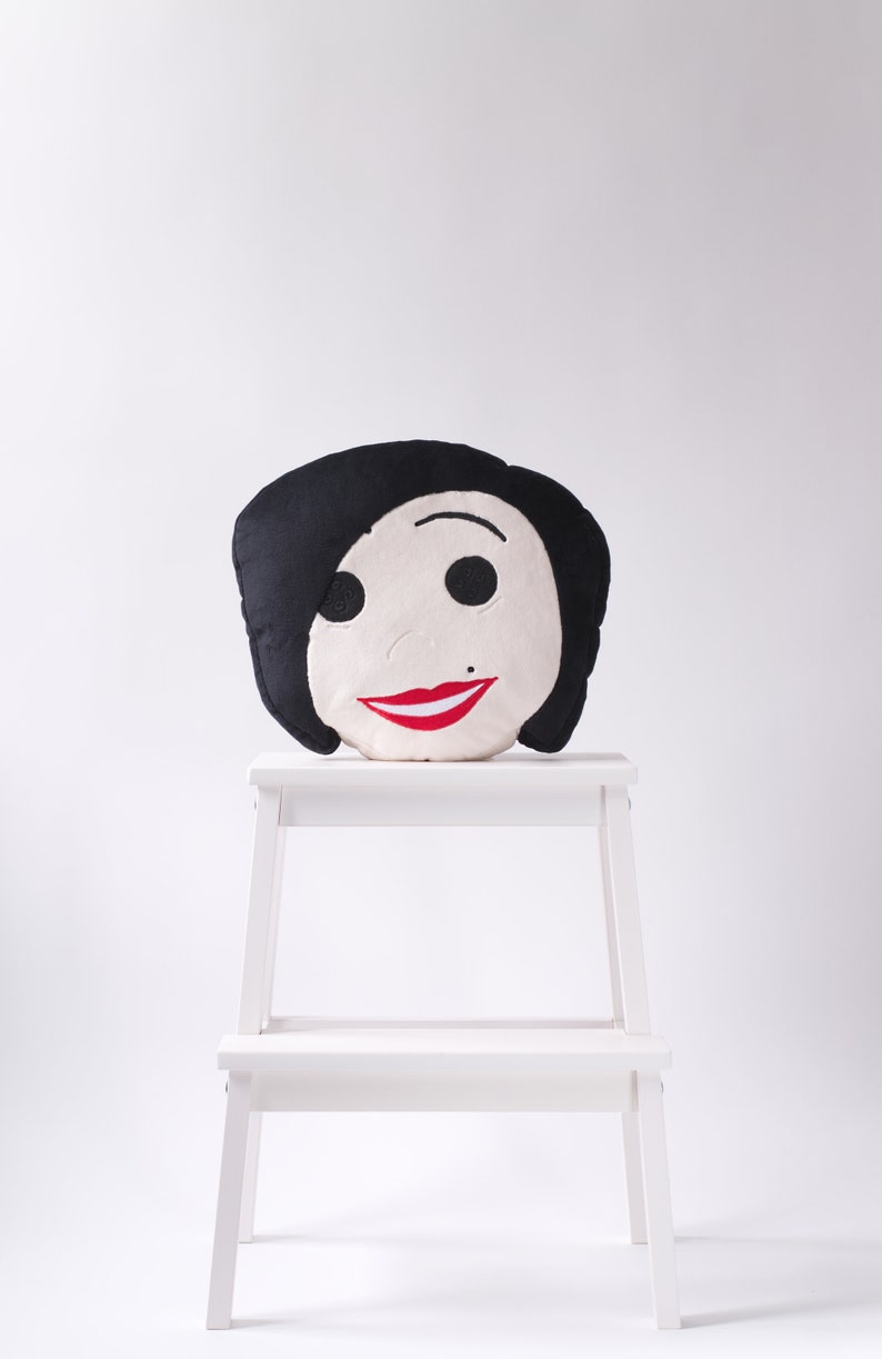 May include: A decorative pillow shaped like a face with black hair, black eyes, a small beauty mark, and a red smile. The pillow is placed on a small white step stool against a white background. The pillow's design is simple and cartoon-like.
