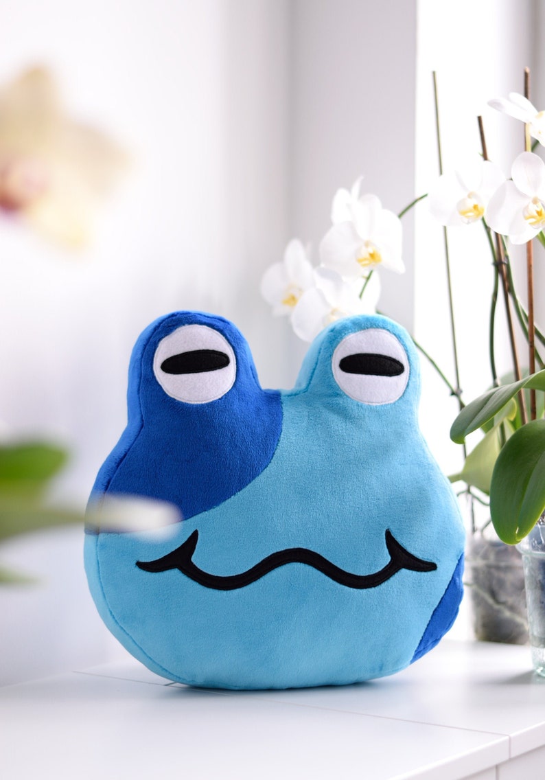 May include: A blue and light blue plush frog with a simple black smile. The frog has white eyes with black pupils.