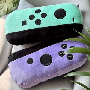 Op de afbeelding: Twee pluche kussens in de vorm van Nintendo Switch Joy-Con controllers. Eén is mintgroen met zwarte knoppen, de andere is paars met zwarte knoppen. De kussens liggen op een grijze ondergrond met groen gebladerte op de achtergrond.