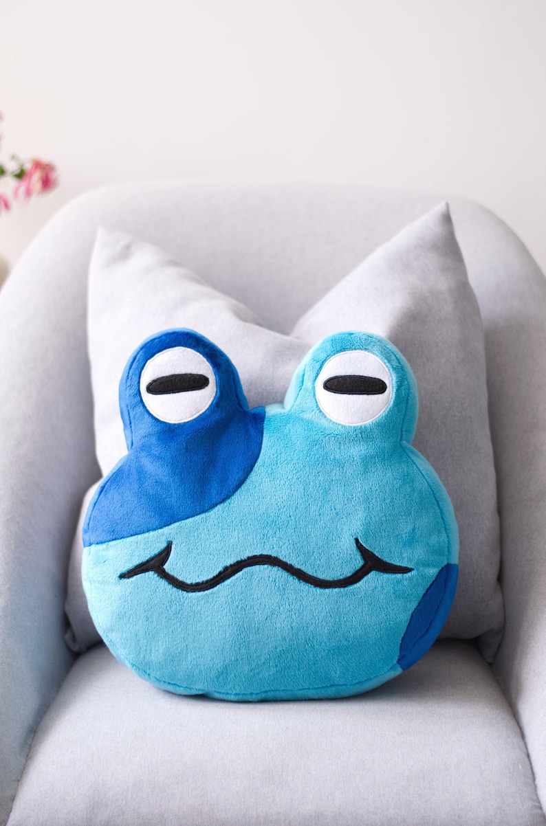 May include: A blue plush frog pillow with a smiling face. The pillow is sitting on a gray couch.