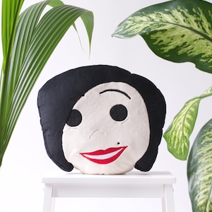 May include: A round decorative pillow featuring a stylized face with black hair, button eyes, and a red smile. The pillow is set against a white background with green foliage.