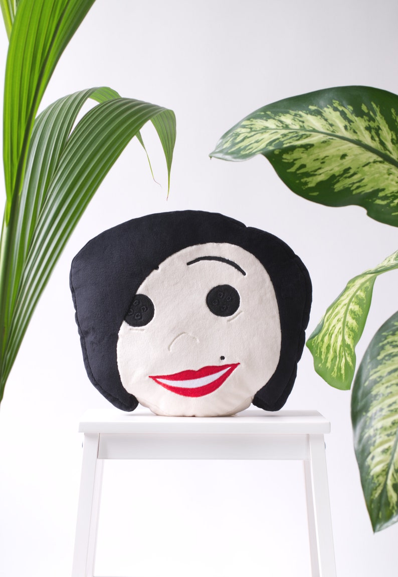 May include: A round decorative pillow featuring a stylized face with black hair, button eyes, and a red smile. The pillow is set against a white background with green foliage.