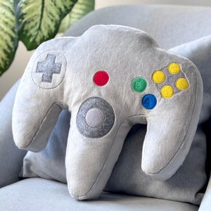 May include: A grey plush pillow shaped like a Nintendo 64 controller. The pillow has a grey fabric with embroidered details in red, green, blue, and yellow.