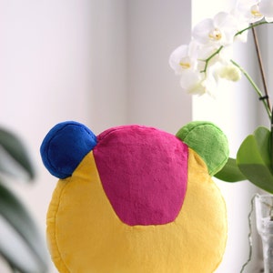 May include: A plush toy shaped like a cartoon character with a yellow body, pink head, blue and green ears.