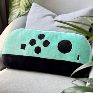 May include: A plush pillow shaped like a teal and black video game controller. The controller has black buttons and a black base. The pillow is resting on a gray couch with green plants in the background.