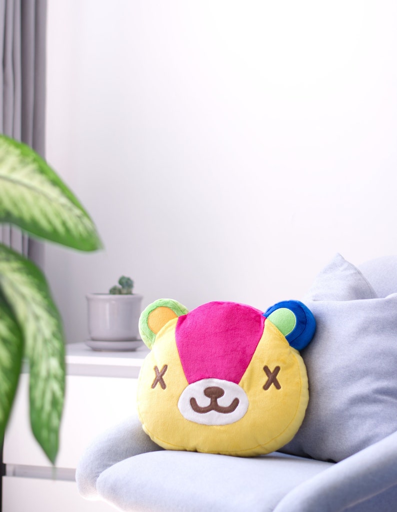 May include: A yellow, pink, blue, and green plush pillow with a cartoon bear face. The bear has brown eyes with an 'X' over each eye, a brown nose, and a brown mouth.
