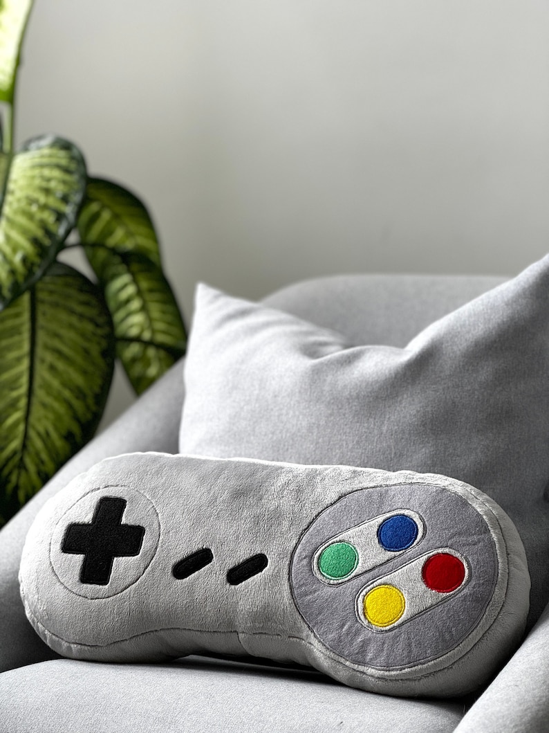 May include: A gray plush pillow shaped like a classic video game controller. The pillow has a black plus sign, four black dots, and four colored buttons: green, blue, red, and yellow.
