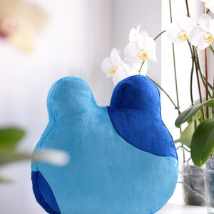 May include: A blue and light blue plush pillow shaped like a bear's head. The pillow is sitting on a white surface in front of a white orchid plant.