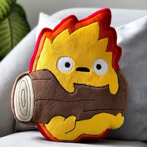 May include: A plush cushion in the shape of a cartoon fire character. The cushion has a yellow flame with red and orange details, white eyes, and a black mouth. It appears to be holding a brown log. The cushion is on a grey surface.