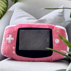 Op de afbeelding: Een roze pluche kussen in de vorm van een draagbare gameconsole. Het kussen heeft een grijs scherm, een wit kruisvormig navigatiepad en roze knoppen. Het kussen ligt op een grijze ondergrond.