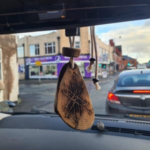 May include: A wooden car air freshener with a carved geometric design, hanging from a rearview mirror. The charm is a light brown colour and is suspended by a beige rope. The background shows a street scene with cars and buildings.