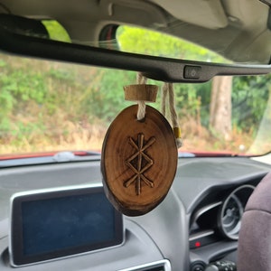 May include: A wooden car charm with a carved Vegvisir symbol, a Norse directional symbol, hanging from a rearview mirror in a car. The charm is attached to a string and a wooden bead.