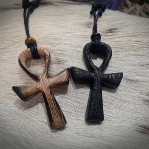 May include: Two wooden Ankh pendants on black cords. One pendant is natural wood with a dark stain, the other is black.