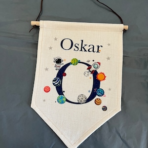 May include: A white canvas banner with a brown leather strap and wooden dowel. The banner features a blue letter "O" surrounded by planets and stars. The name "Oskar" is printed above the letter.