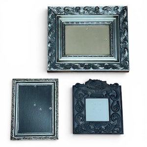 May include: Three empty picture frames: one large rectangular frame with a silver finish and ornate leaf detailing, one vertical rectangular frame with a silver beaded edge, and one square frame with a dark finish and intricate floral carvings.
