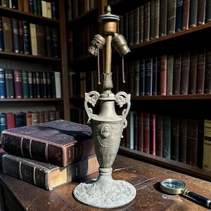 May include: An antique brass lamp with a detailed urn-shaped base and two light sockets. The lamp sits on a wooden table next to a stack of old books and a magnifying glass. The background features a library with many books.