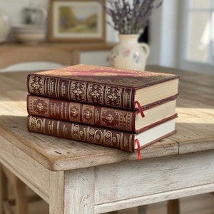 May include: Three antique books stacked on a rustic wooden table. The books have burgundy leather covers with gold embossed designs and red ribbon bookmarks. The table is light-colored with visible wood grain.