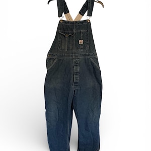 May include: Dark blue denim overalls hanging on a black hanger. The overalls have a chest pocket, button closures, and adjustable straps. A small patch is visible on the chest pocket. The overalls are a classic workwear style.