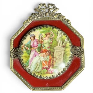 May include: An octagonal decorative wall plaque featuring a central circular porcelain insert with a colorful painting of three figures in a garden setting. The insert is framed by a deep red border and an ornate gold-tone metal frame with a bow top.