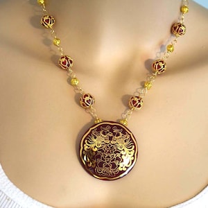 May include: A gold necklace with a large, round pendant featuring a red enamel design of two phoenixes and a flower. The necklace is made of gold chain and red beads.