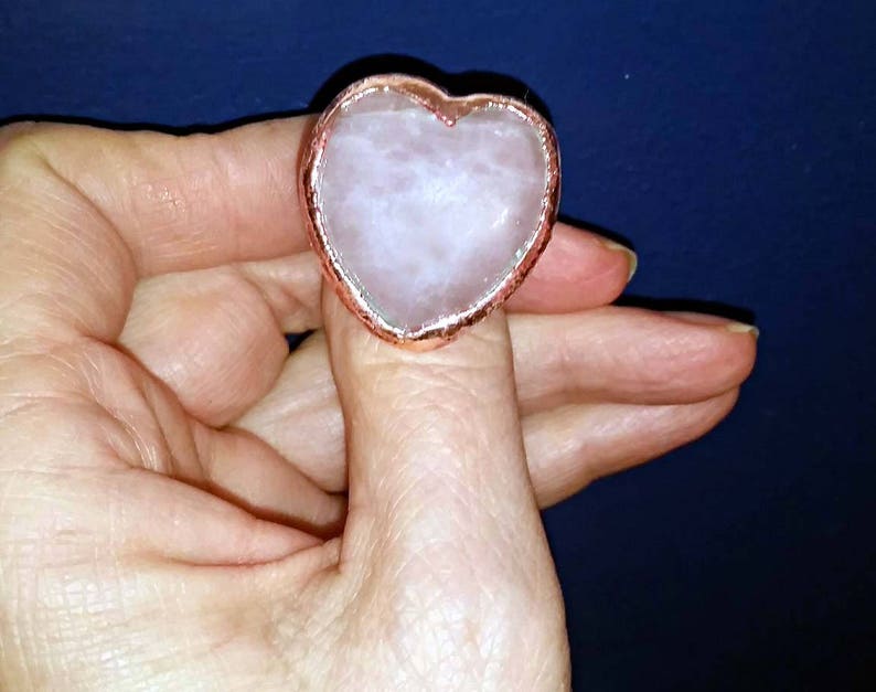 Pode incluir: Um anel em forma de cora&ccedil;&atilde;o com uma pedra de quartzo rosa engastada em uma moldura de metal de cor cobre.