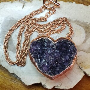 May include: A rose gold necklace with a large amethyst heart pendant. The heart is filled with small amethyst crystals and has a rose gold border.