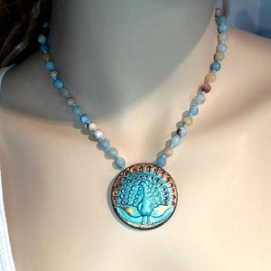 May include: A turquoise blue necklace with a round pendant featuring a peacock design. The pendant has gold accents and red accents around the peacock's tail feathers. The necklace is made of light blue and brown beads.