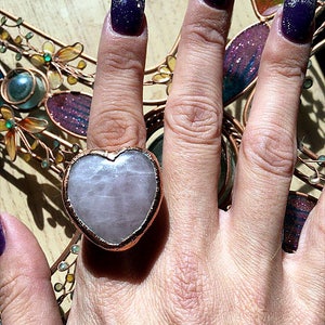 Pode incluir: Um anel em forma de cora&ccedil;&atilde;o com uma pedra rosa clara engastada em uma banda de metal de tom cobre. O anel &eacute; usado no dedo indicador de uma m&atilde;o com esmalte de unha roxo escuro.