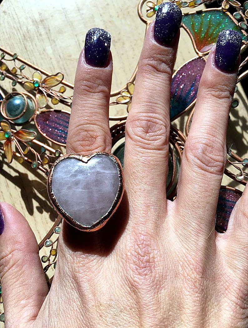 Pode incluir: Um anel em forma de cora&ccedil;&atilde;o com uma pedra rosa clara engastada em uma banda de metal de tom cobre. O anel &eacute; usado no dedo indicador de uma m&atilde;o com esmalte de unha roxo escuro.