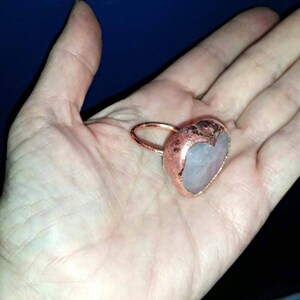 Pode incluir: Um anel de cobre com uma pedra de quartzo rosa em forma de cora&ccedil;&atilde;o. O anel &eacute; feito &agrave; m&atilde;o e tem um acabamento r&uacute;stico e martelado.