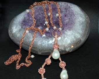 Pode incluir: Um colar de corrente de cobre com um pingente com um design de ramo com três gotas de pérolas. O colar é exibido em uma pedra de ágata roxa e branca.