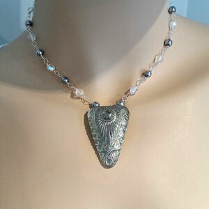 May include: Silver-toned necklace featuring a pendant with an ornate, engraved design. The pendant is a stylized triangle shape. The necklace chain is strung with small, dark gray beads and clear, faceted beads.