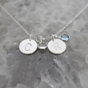 May include: A silver necklace with three pendants. Two round pendants are engraved with the letters "C" and "R". A small, clear crystal and a blue gemstone are also attached to the chain. The necklace is displayed on a gray surface.