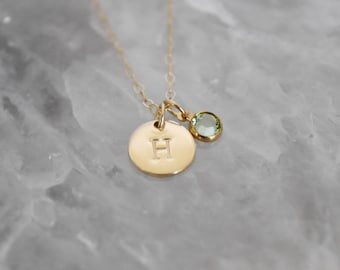May include: A gold-colored necklace with a round pendant engraved with the letter "H" and a small, round, light green gemstone charm. The necklace is displayed on a light gray, marbled surface.