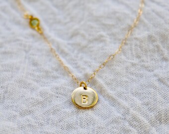 May include: A delicate gold-colored necklace with a small, round pendant featuring the letter "B". The necklace has a fine chain and a small green gemstone accent. The jewelry is displayed on a white, textured surface.