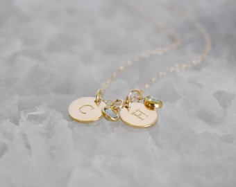 May include: A delicate gold necklace featuring two circular pendants, each engraved with a letter. One pendant has the letter "C" and the other has the letter "H". The necklace also includes two small gemstone charms.