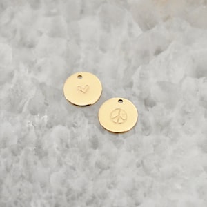 May include: Two small, gold-colored, round charms. One charm features a heart symbol, and the other displays a peace sign. Each charm has a small hole at the top for stringing. The charms are displayed on a light-colored, textured surface.