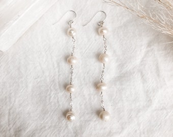 Puede incluir: Pendientes colgantes con cuatro perlas blanquecinas suspendidas de una cadena de plata. Los pendientes tienen un diseño de gancho simple y se muestran sobre una superficie blanca texturizada. Las perlas tienen una forma natural e irregular.