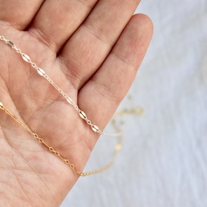 May include: Close-up of two delicate necklaces, one gold and one silver, each featuring a chain of small, elongated oval links. The necklaces are held in a hand against a soft, white background, showcasing the jewelry's fine details.