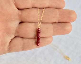 May include: A gold necklace with a vertical bar pendant. The pendant is made of small, deep red gemstones. The necklace is shown against a plain background, emphasizing the jewelry's design and colour.