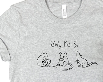 May include: A gray t-shirt with the text "aw, rats." and a simple line drawing of three rats. The shirt is displayed on a white background with a pair of blue jeans and sunflowers.