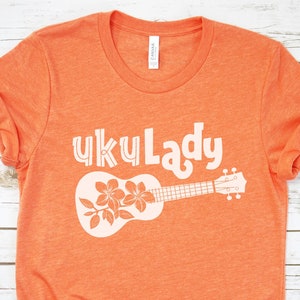 May include: Orange t-shirt with white text that says "Uku Lady" and a white ukulele with flowers on it.