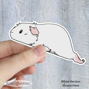 May include: White sticker of a cartoon guinea pig with pink ears and a white body. The sticker is being held by a hand.